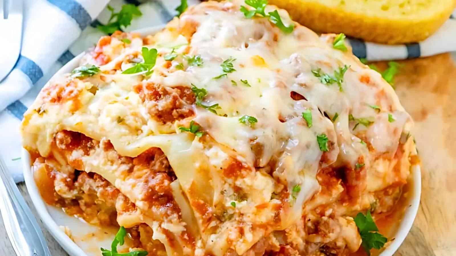 A close-up view of a slice of cheesy lasagna on a plate, topped with melted cheese and garnished with chopped parsley. A piece of garlic bread and a checkered napkin are visible in the background.