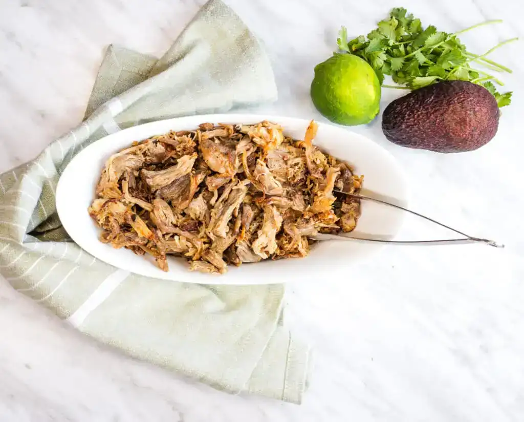 Shredded pork served on a white oval dish, accompanied by a green lime, a whole avocado, and a bunch of fresh cilantro on the side. The dish is placed atop a green and white striped cloth on a marble surface. Silver tongs are resting on the dish.