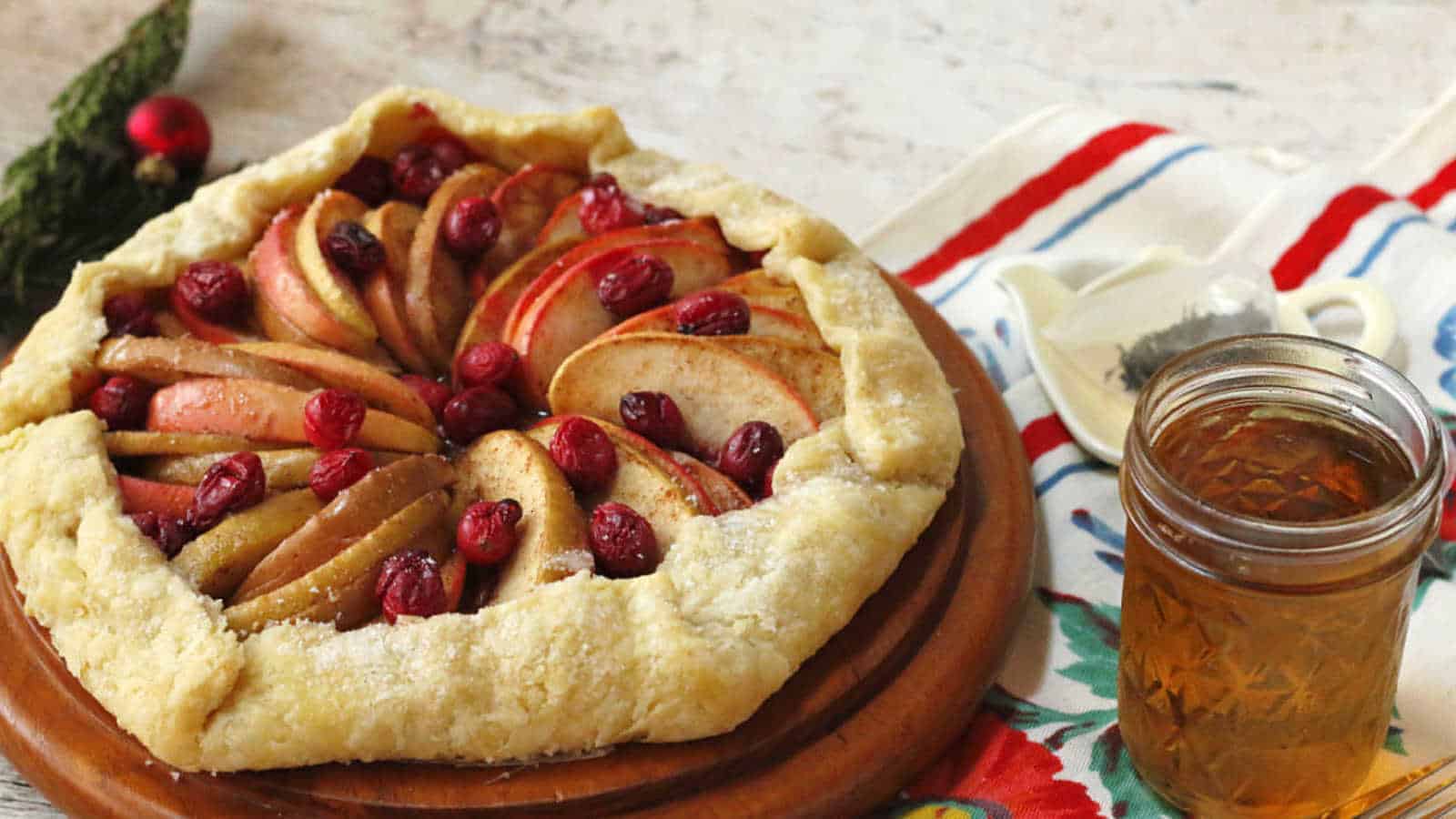 A rustic apple cranberry galette sits on a wooden board. The golden-brown crust encases sliced apples and cranberries arranged in a circular pattern. A jar of honey is to the side, along with a colorful cloth napkin, creating a warm and inviting setting.