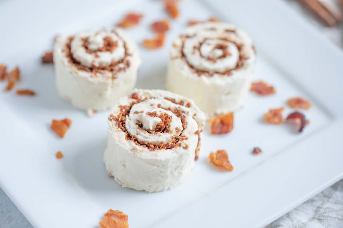 Three cinnamon rolls topped with frosting are placed on a white square plate. Small pieces of bacon are scattered around the rolls on the plate. The rolls have a swirled appearance with a rich brown cinnamon filling.