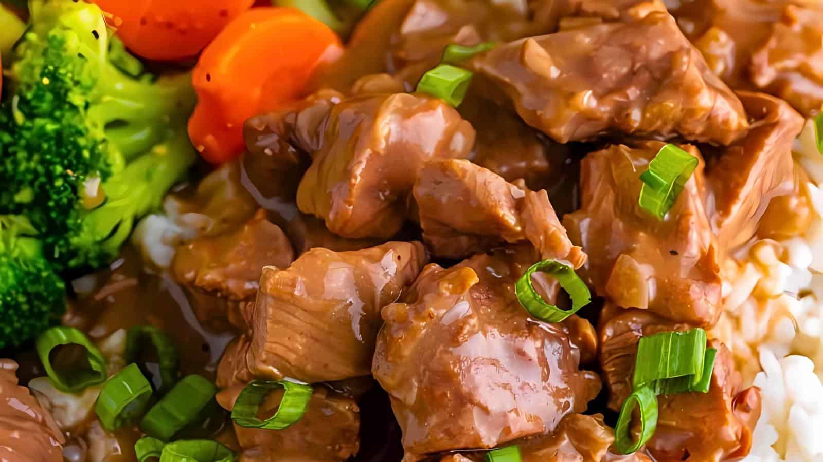 Close-up of a plate featuring tender pieces of beef stew garnished with sliced green onions. The beef is served atop a bed of rice, with steamed broccoli and sliced carrots on the side, all drenched in a savory brown sauce.