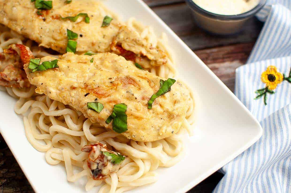 A rectangular white plate holds a serving of creamy fettuccine pasta topped with two pieces of golden, herb-seasoned chicken breast. Fresh basil leaves are scattered on top. A white striped napkin with a yellow flower is to the right, and a small bowl of sauce is in the background.