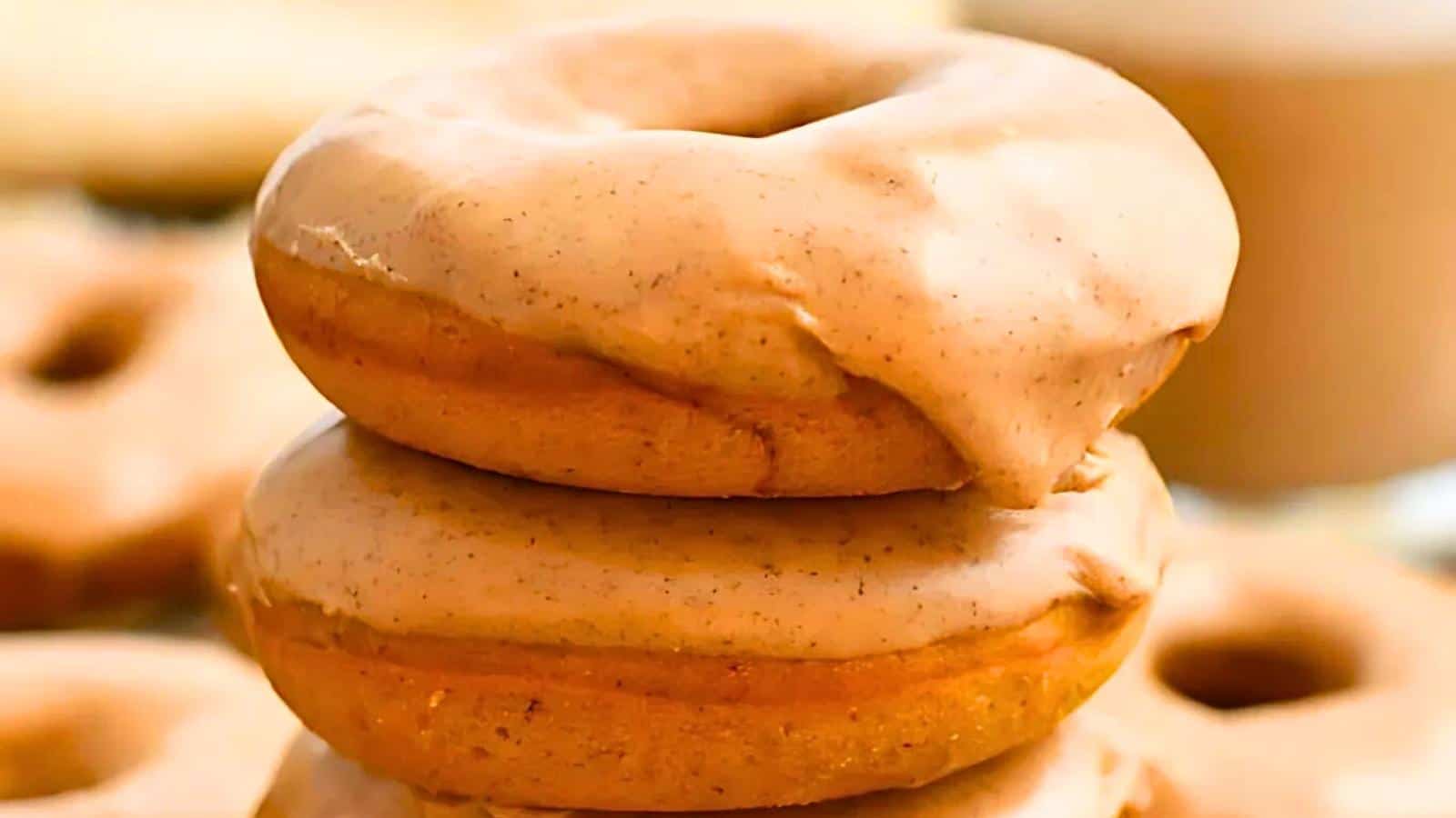 A close-up image of two glazed donuts stacked on top of each other. The donuts have a light brown glaze that appears creamy and smooth, with a slightly textured surface. The background is softly blurred, highlighting more donuts and a cup, possibly of coffee.