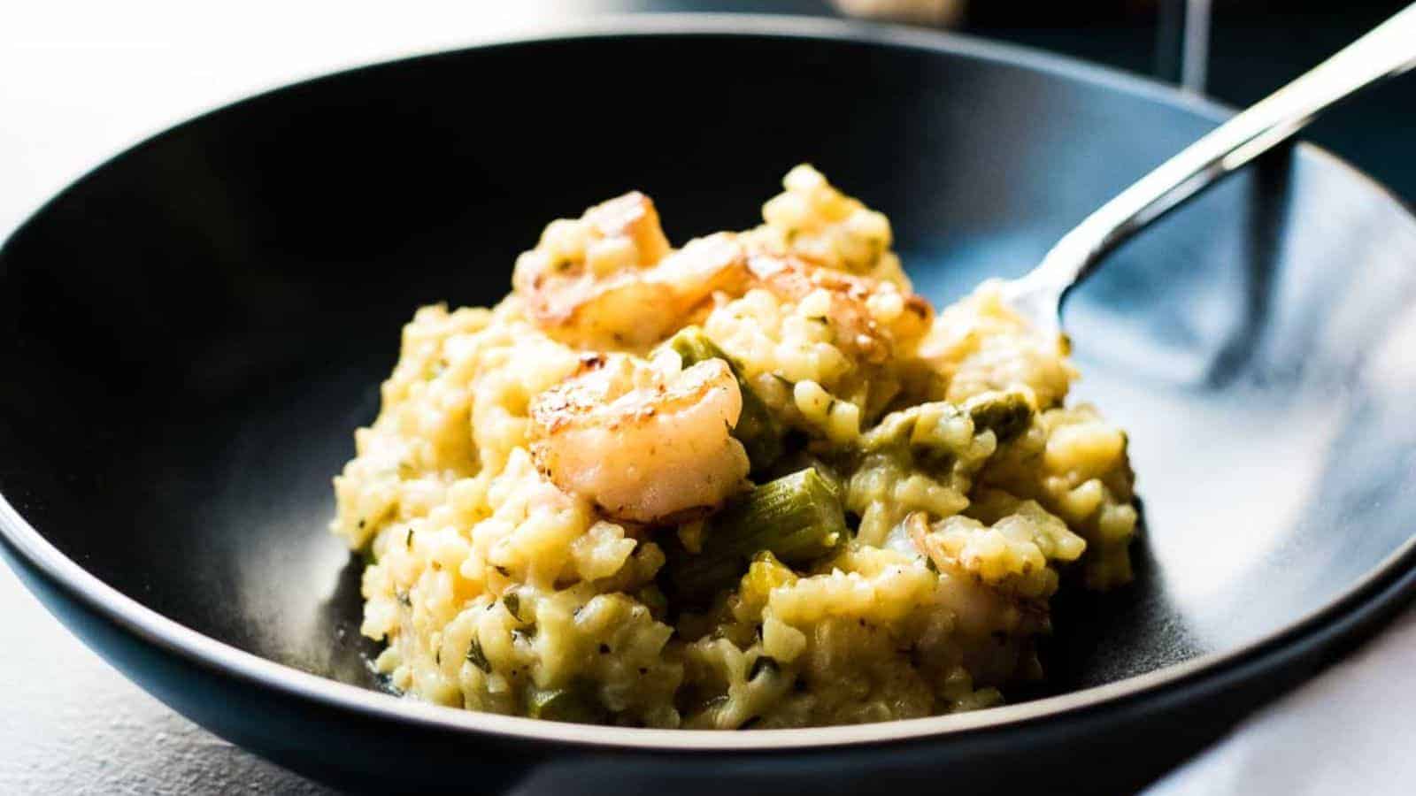 A close-up of a black bowl containing creamy risotto mixed with shrimp and green vegetables. A fork is partially visible, resting on the edge of the bowl, ready to scoop up a bite. The dish has a golden hue with visible seasonings.
