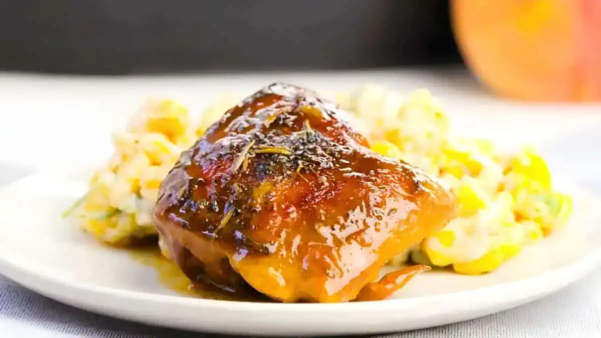 A glazed piece of salmon with a caramelized surface sits atop a white plate, accompanied by a side of creamy corn salad. The dish is garnished with herbs and presented on a white tablecloth with a blurred background, emphasizing the vibrant colors and textures of the food.