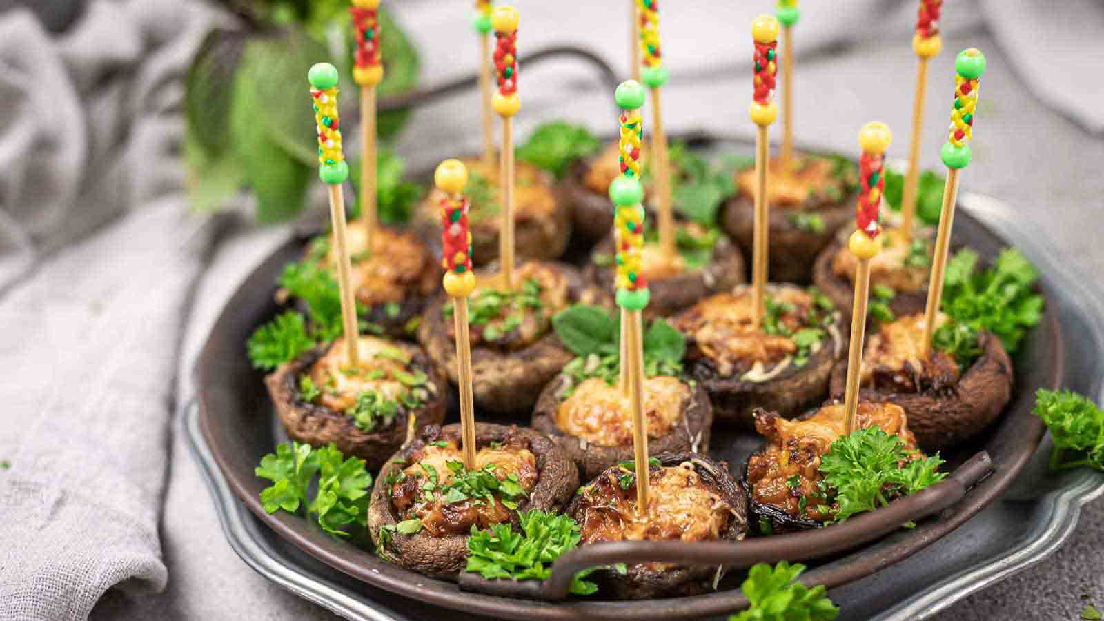 A plate of stuffed mushrooms garnished with chopped herbs. Each mushroom has a colorful toothpick inserted, making them easy to pick up. The plate is surrounded by fresh parsley and a green leafy plant is visible in the background.