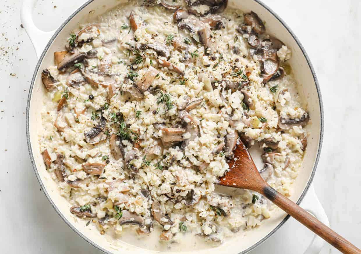A creamy mushroom risotto dish is shown in a white pan. The risotto is mixed with sliced mushrooms and sprinkled with fresh herbs. A wooden spoon rests inside the pan, indicating the dish is freshly stirred.