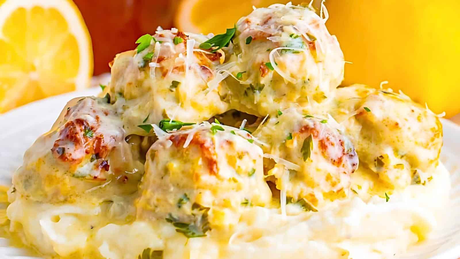 A plate of creamy chicken meatballs topped with melted cheese and fresh parsley. The meatballs rest on a bed of mashed potatoes, with visible lemon slices and a yellow-orange background. The dish is garnished with grated parmesan and has a golden-brown crust.