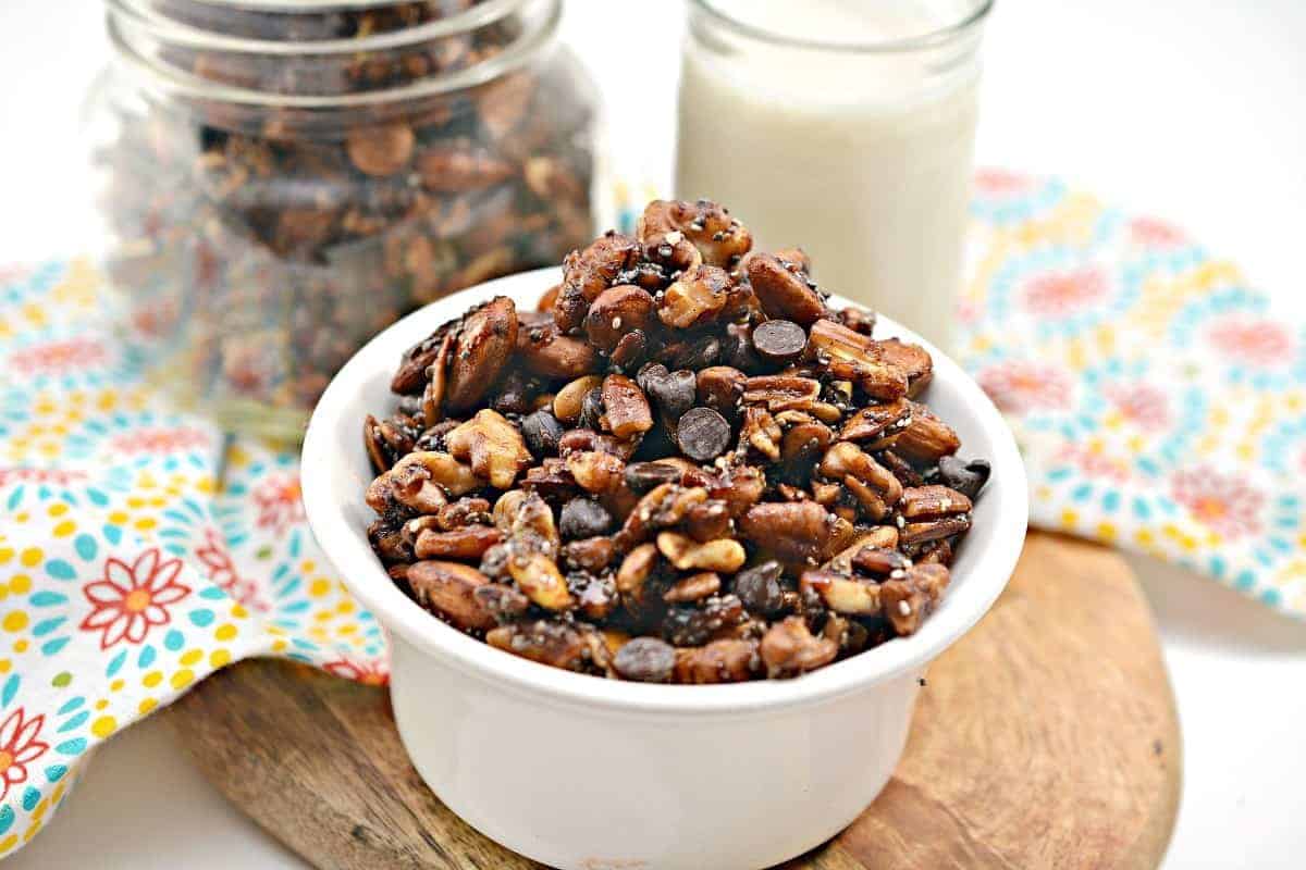 A white bowl filled with a mixture of nuts and chocolate chips is placed on a wooden surface. Behind the bowl, there's a mason jar containing more of the same mixture, and a glass of milk. A colorful, patterned cloth is positioned beside the bowl and jar.