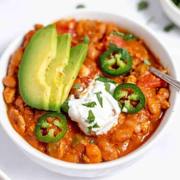 A bowl of chili topped with sliced avocado, jalapeño, sour cream, and chopped herbs, with a spoon on the side.