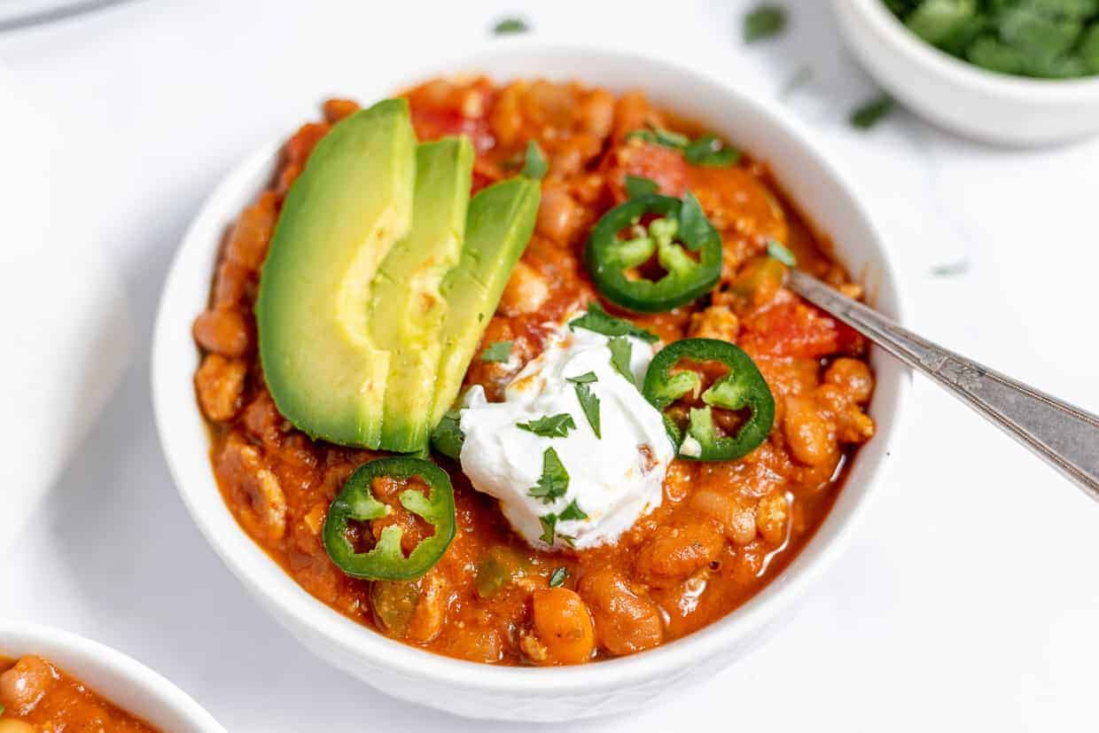 A bowl of chili topped with sliced avocado, jalapeño slices, and a dollop of sour cream. A spoon rests in the bowl, ready to scoop up the spicy, hearty meal. In the background, a small bowl of chopped herbs or green onions is partially visible.