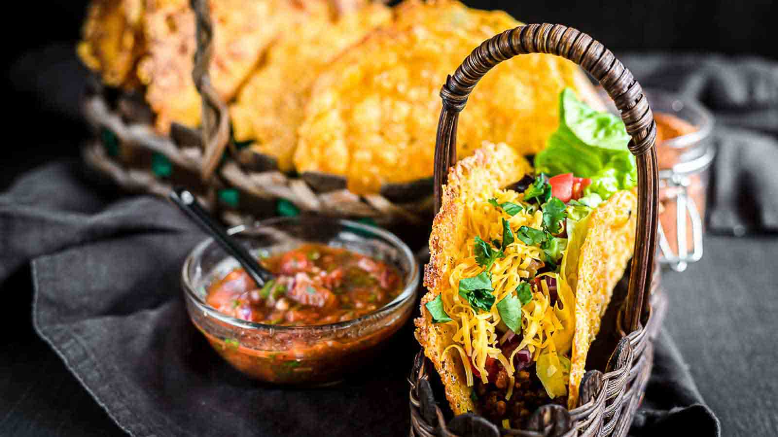 A wicker basket contains a taco filled with ground meat, shredded cheese, lettuce, and diced tomatoes. A small bowl of salsa with a spoon sits beside the basket. In the background, more tacos are arranged in another wicker basket on a dark tablecloth.