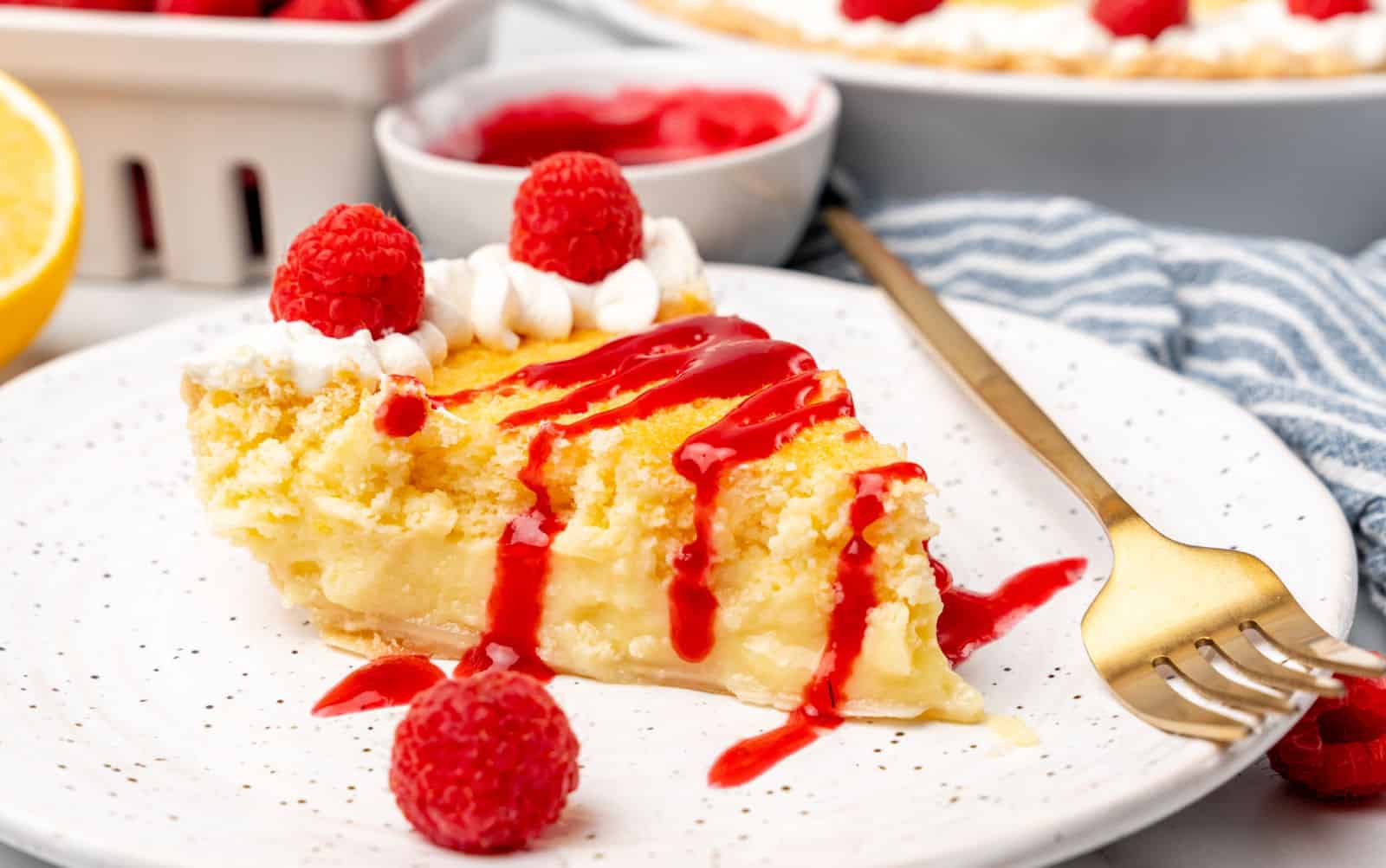 A close-up of a slice of lemon pie on a white speckled plate. The pie is topped with whipped cream, fresh raspberries, and a drizzle of red sauce. A gold fork rests on the plate, and additional raspberries and pie are visible in the background.