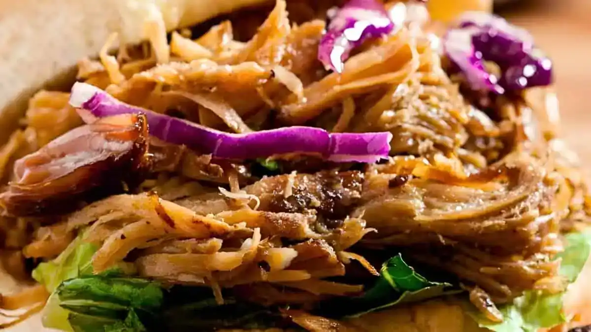Close-up of a pulled pork sandwich with leafy greens and red cabbage slaw. The tender, shredded pork appears juicy and flavorful, while the cabbage adds a pop of color and crunch to the sandwich. The ingredients are layered in a bun.