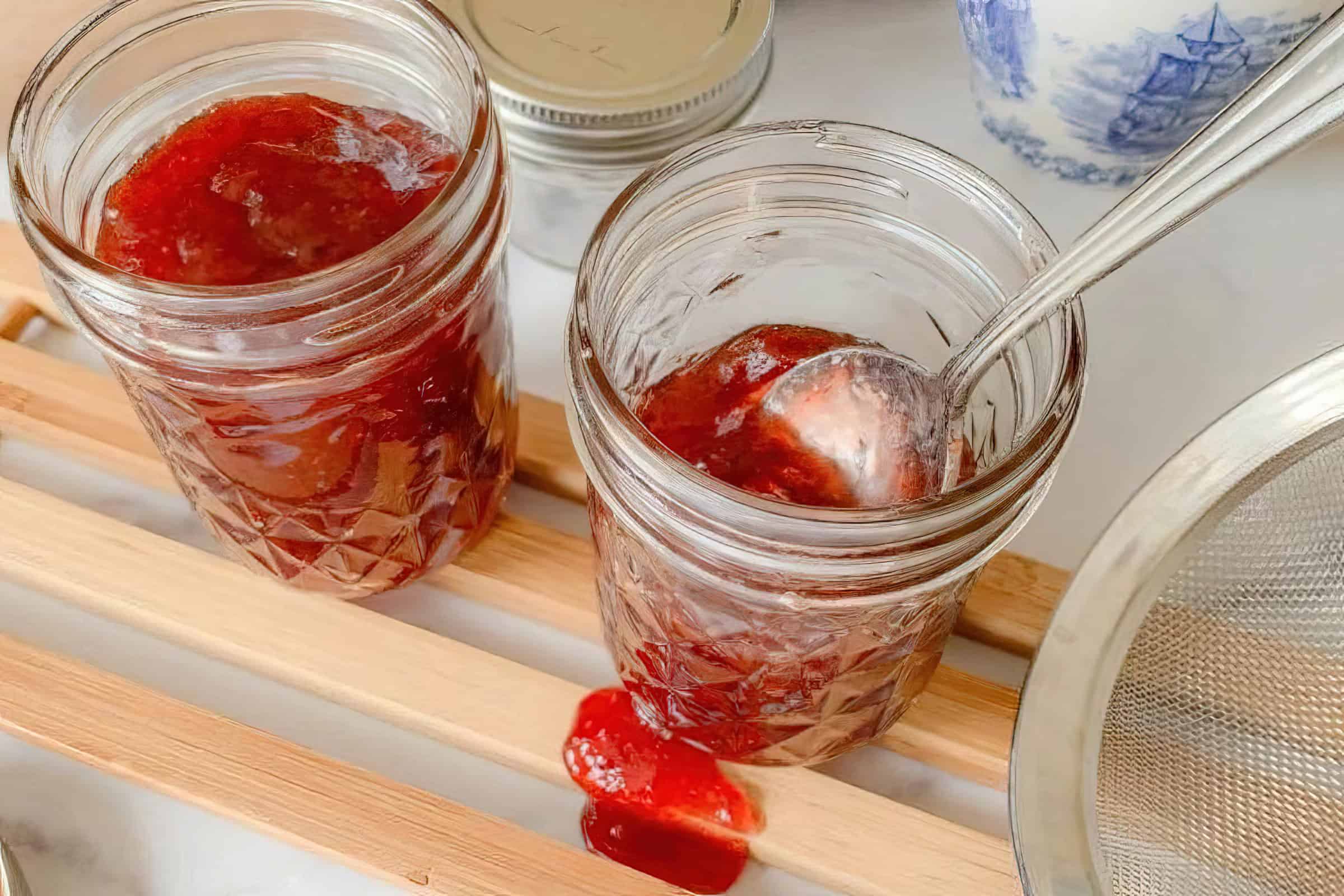 Two open glass jars filled with strawberry jam, one with a spoon inside. The jars are placed on a wooden rack, with a small blob of jam on the rack. A metal sieve and a partially visible ceramic container are in the background.