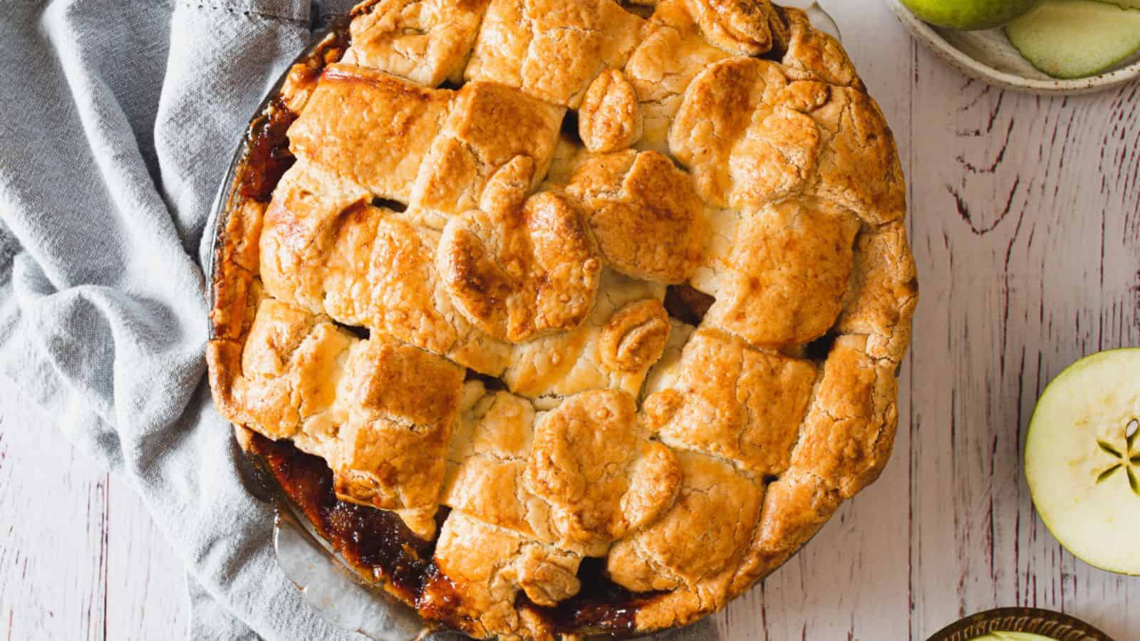 A freshly baked apple pie with a golden-brown lattice crust sits on a white wooden surface. A light blue cloth is partially visible next to the pie, and a halved green apple with seeds exposed is seen to the right of the pie.