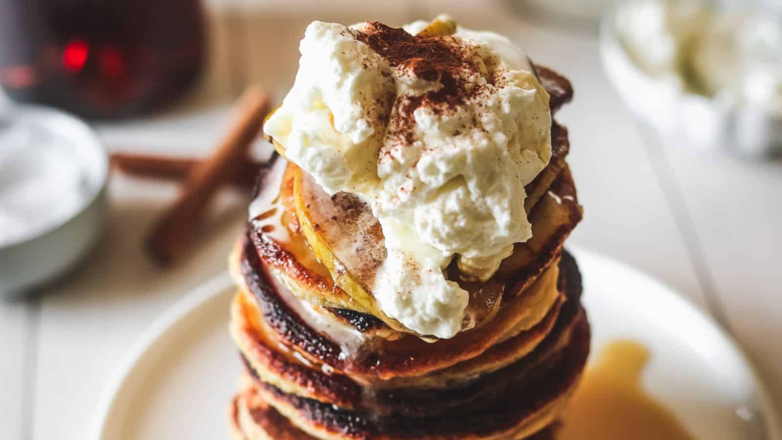 A stack of pancakes on a white plate, topped with a generous dollop of whipped cream and a sprinkle of cinnamon. The pancakes are drizzled with maple syrup, and there are cinnamon sticks and a small bowl in the blurred background.