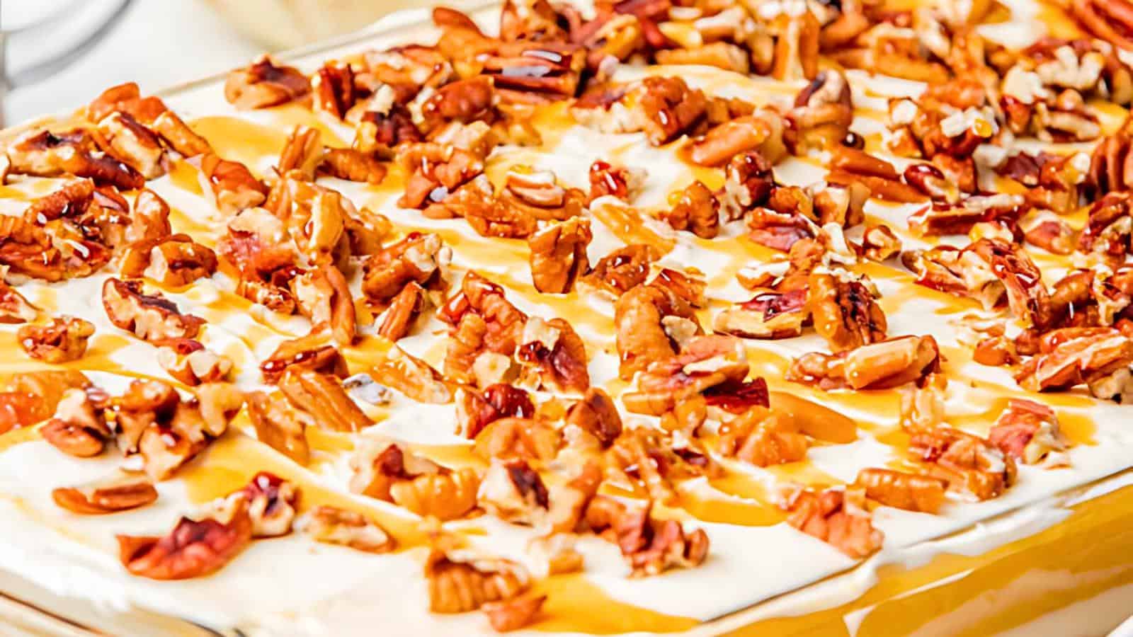 A close-up of a dessert topped with a generous layer of chopped pecans and drizzled with a light caramel sauce. The dessert appears to have a creamy frosting or icing underneath the pecans, creating a rich and indulgent appearance.