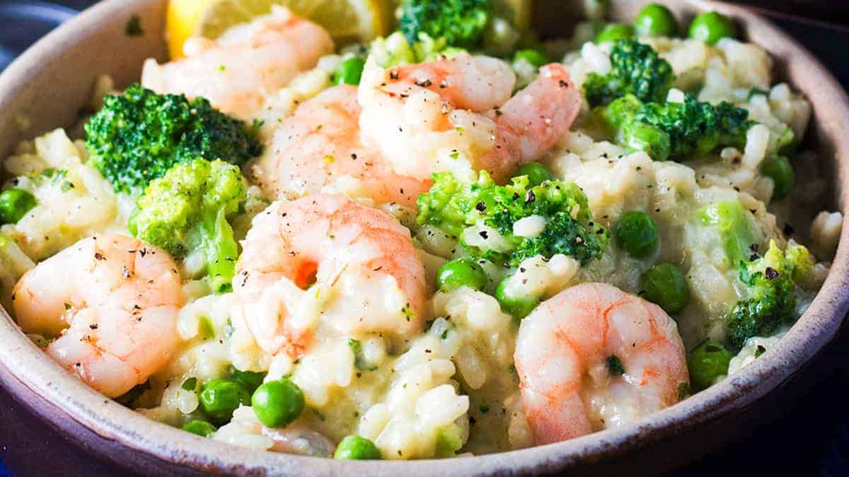 A bowl of shrimp and vegetable risotto. The dish includes plump shrimp, bright green broccoli florets, and peas in a creamy rice base. There are lemon wedges at the edge of the bowl, and the meal is garnished with a sprinkle of cracked black pepper.