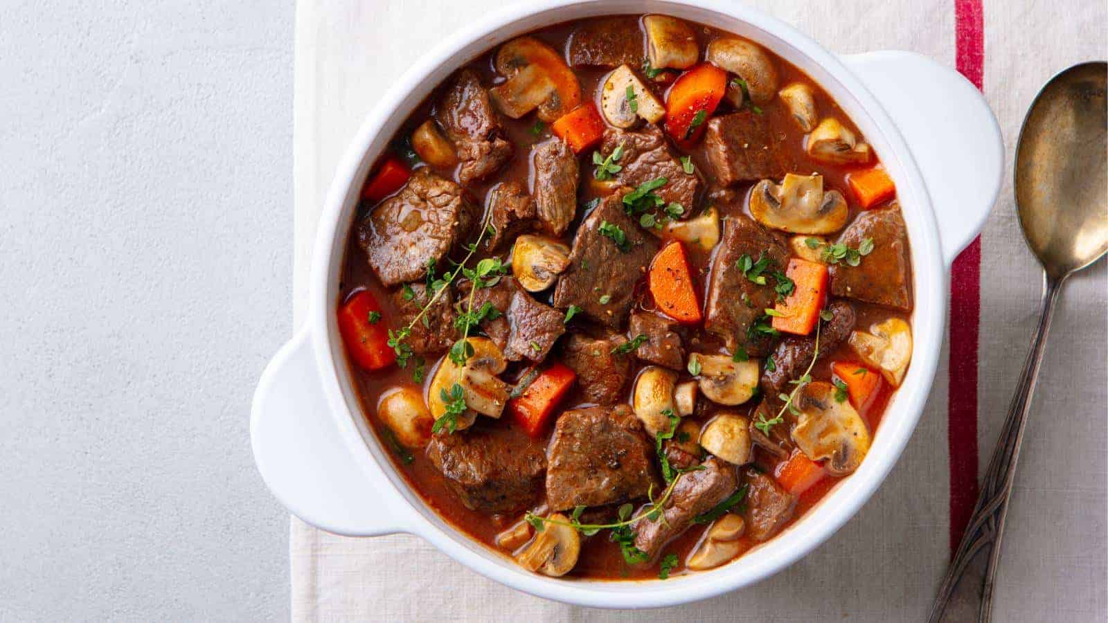 A white ceramic dish filled with beef stew featuring chunks of beef, sliced mushrooms, and carrot pieces in a rich brown sauce. The dish is garnished with fresh herbs and placed on a white cloth next to a brass spoon.