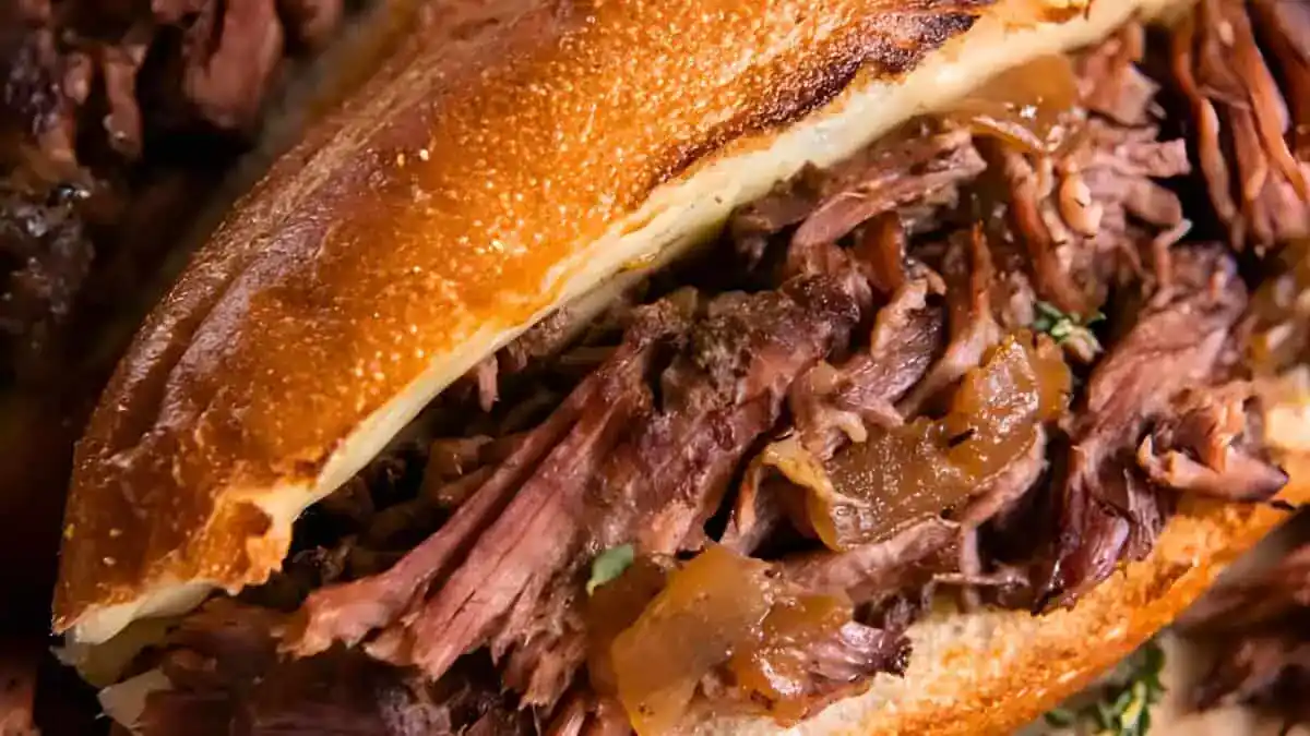 A close-up of a savory roast beef sandwich. The sandwich features tender, shredded roast beef with visible herbs and caramelized onions, all nestled inside a crispy, toasted bun. The textures and colors highlight the deliciousness of the meat and bread.