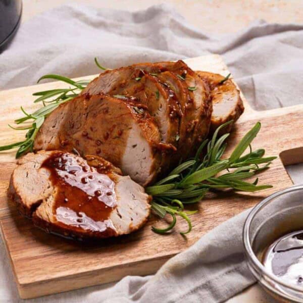 Sliced roasted pork loin with sauce and fresh rosemary on a wooden cutting board, with a bowl of extra sauce on the side.