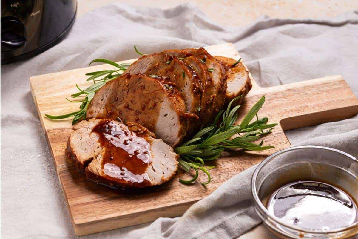 A seasoned, roasted pork loin is sliced and displayed on a wooden cutting board, garnished with fresh rosemary. A small bowl of glaze sits nearby, and a slow cooker is partially visible in the background on a light cloth-covered surface.