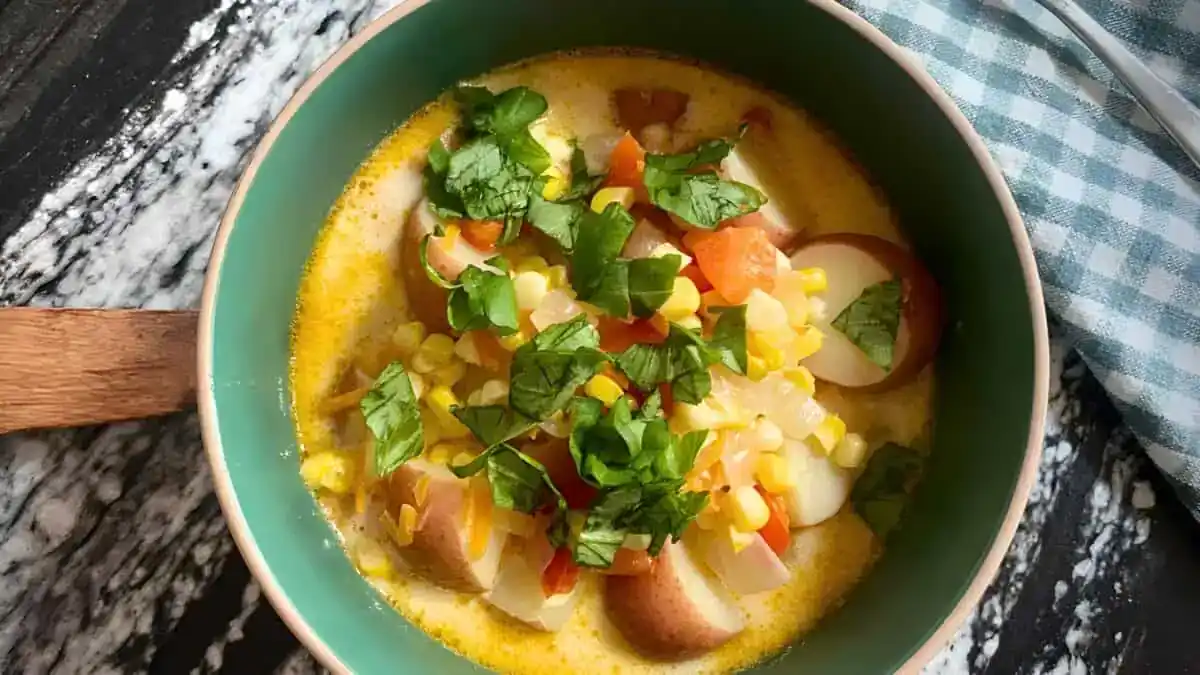 A bowl of creamy chowder featuring diced potatoes, corn kernels, chopped carrots, and onions, garnished with fresh cilantro. The bowl has a handle, and a checkered cloth is partially visible in the background on a dark marbled surface.