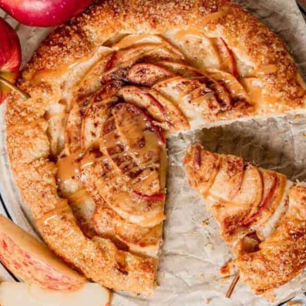 A rustic apple galette on parchment paper rests on a cooling rack, with fresh apples and cinnamon sticks arranged around it.