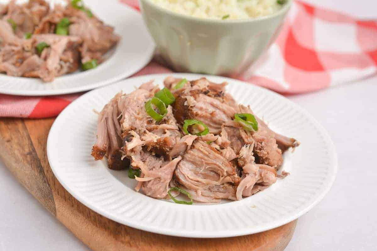 A white plate filled with shredded pork garnished with green onions sits on a wooden board. In the background, a bowl of rice and another plate with a similar dish can be seen, along with a red and white checkered cloth.