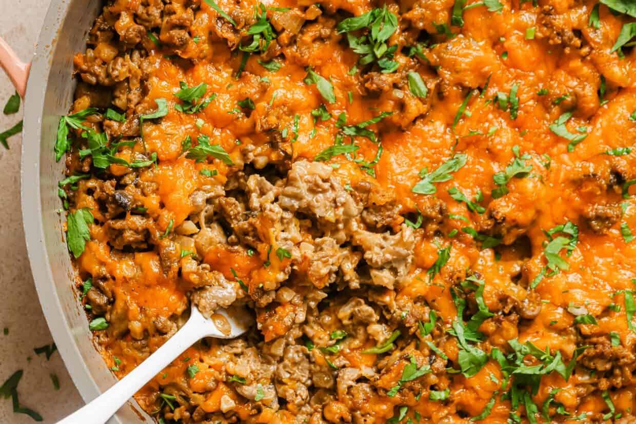 A close-up view of a cheesy ground beef skillet topped with melted cheese and chopped parsley. A spoon is partially submerged in the mixture, revealing the cooked ground beef underneath.