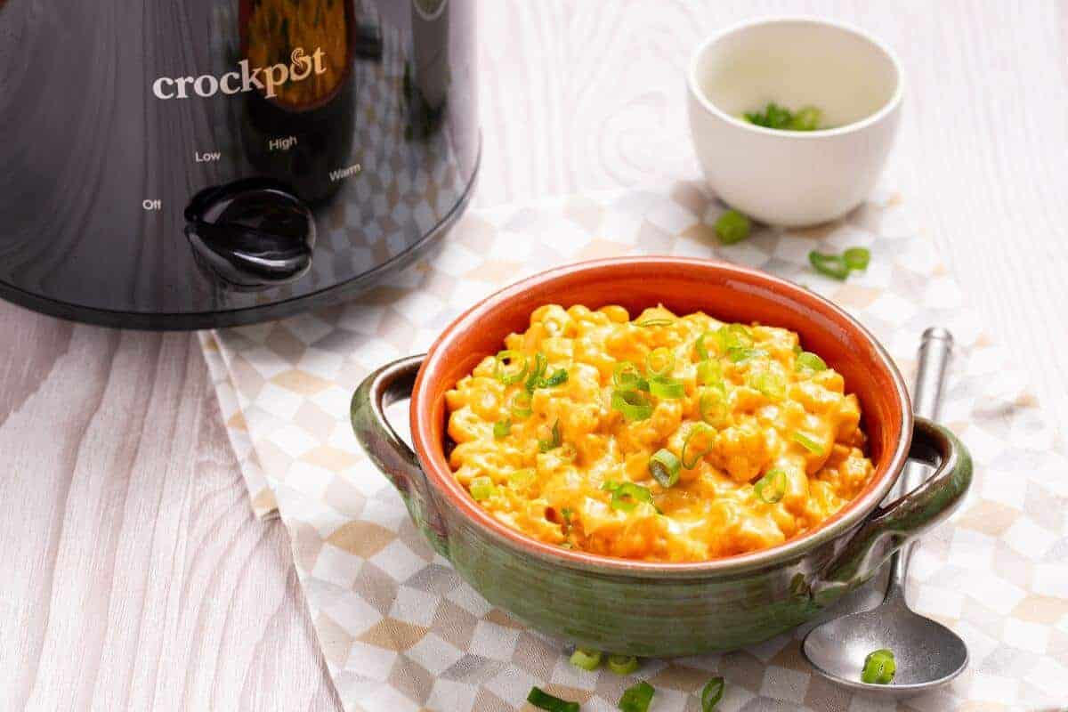 A green bowl filled with cheesy macaroni and cheese garnished with chopped green onions, next to a spoon on a checkered cloth. A small white bowl with more green onions and a crockpot are visible in the background.