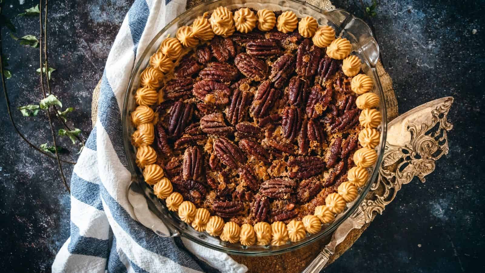 A pecan pie topped with whole pecans and a scalloped border of piped cream sits on a decorative serving platter. It is partially wrapped in a white and navy striped kitchen towel, with a pie server visible beside it. The background is a dark, textured surface.