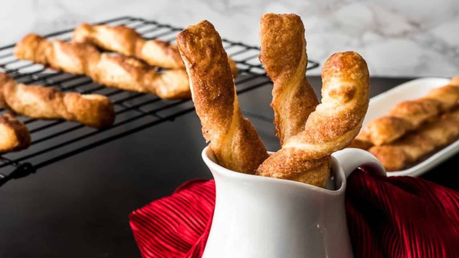 A white pitcher holds three golden-brown, twisted breadsticks, with more breadsticks in the background on a cooling rack and a white plate. A red cloth is draped under the pitcher, adding a pop of color to the scene. The surface and backdrop are dark.