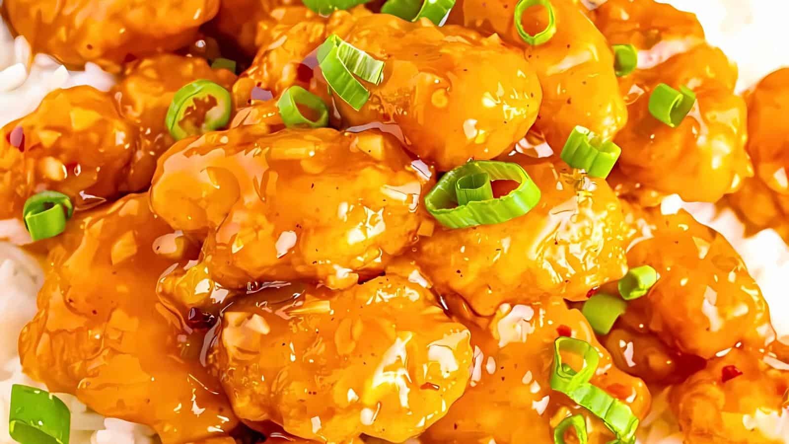 Close-up of a plate of orange chicken, featuring crispy, golden-brown chicken pieces coated in a glistening, tangy orange sauce. The dish is garnished with sliced green onions and served on a bed of white rice.