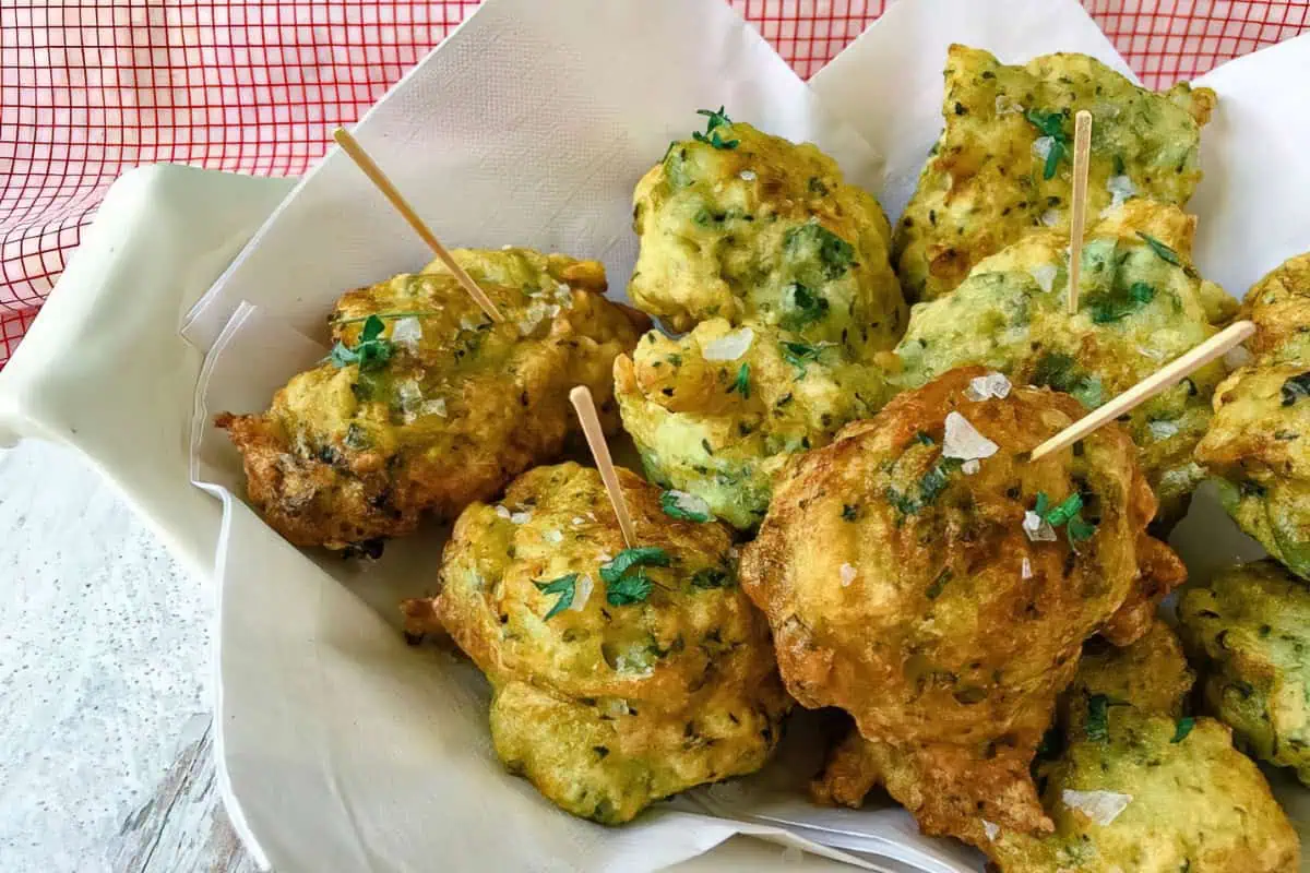A plate of freshly fried, golden brown fritters garnished with chopped herbs and coarse salt. Some fritters have toothpicks inserted for easy handling. They are served on white paper placed in a basket, with a checkered red and white background.