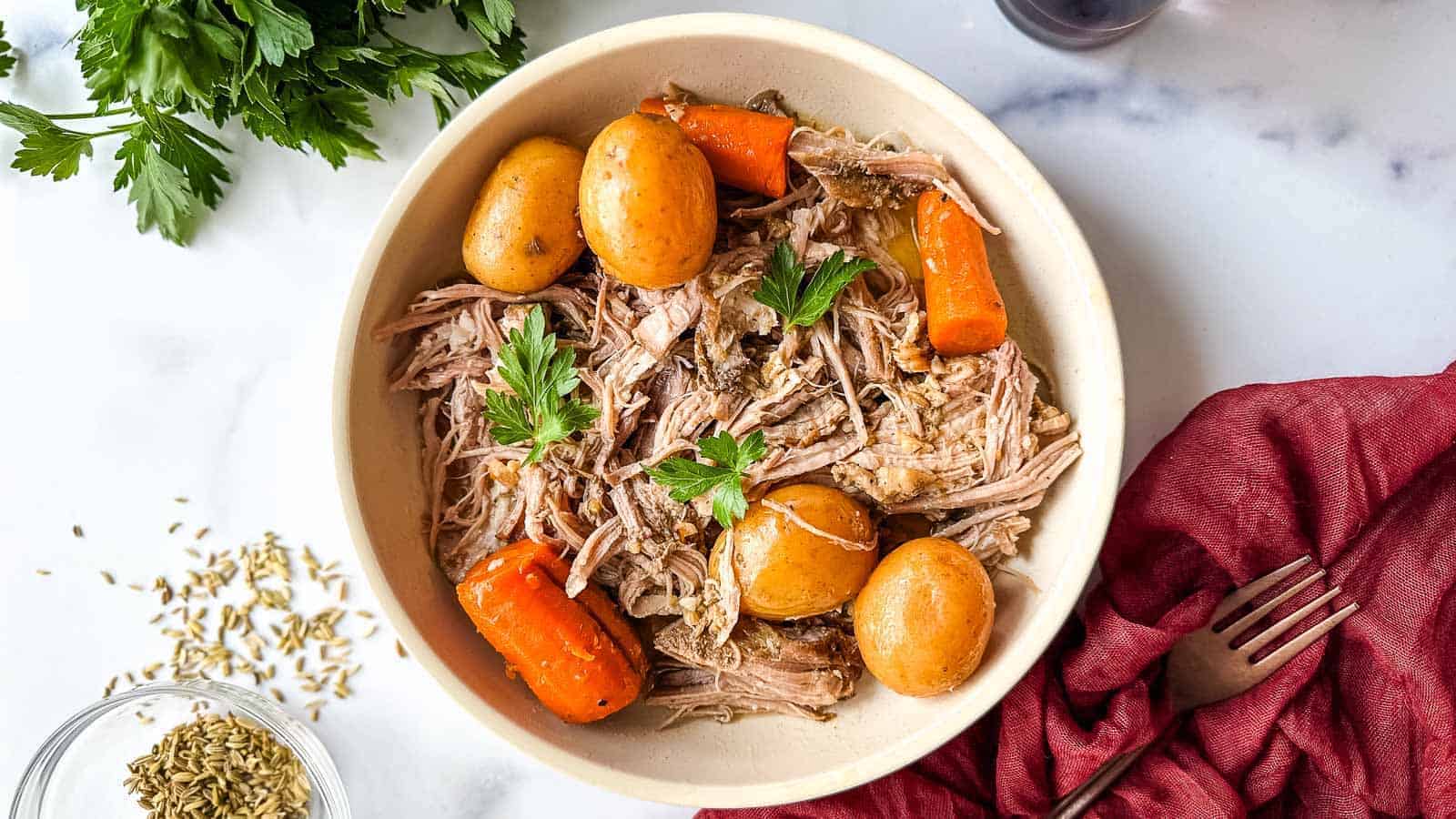A bowl filled with shredded pork, whole baby potatoes, and large carrot chunks, garnished with parsley. A red cloth and fork are placed beside the bowl. There are also some fennel seeds and parsley leaves visible on the side.
