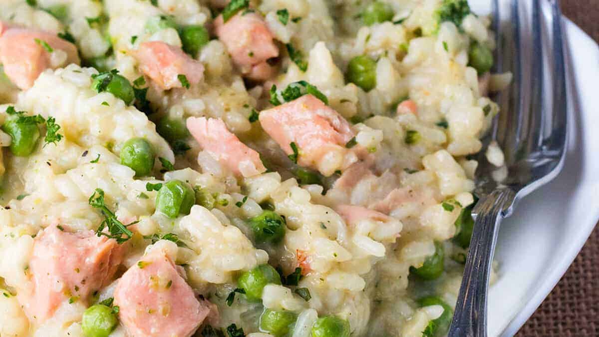 A close-up of a creamy rice dish featuring chunks of pink salmon, green peas, and finely chopped herbs. The dish is garnished with parsley and presented on a white plate with a set of silver forks resting on the side.