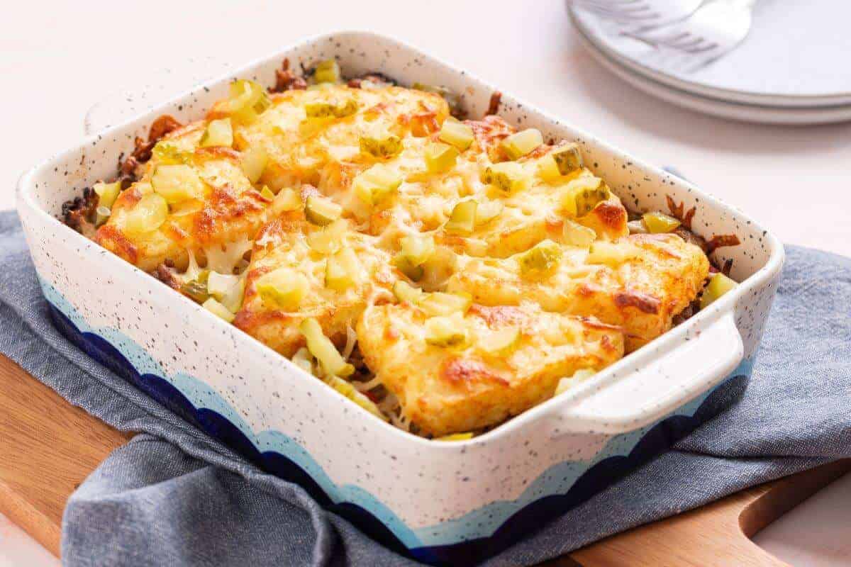 A freshly baked casserole dish filled with a golden-brown tater tot casserole topped with melted cheese and chopped green peppers. The dish sits on a blue cloth on a wooden surface with plates and utensils in the background.