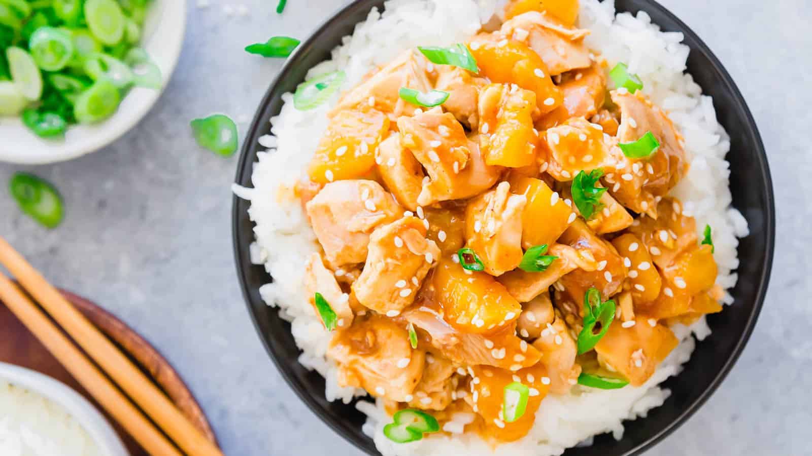 A bowl of white rice topped with chunks of chicken and small pineapple pieces, all coated in a glistening orange sauce. The dish is garnished with sesame seeds and chopped green onions. To the side, there are chopsticks and a small bowl of sliced green onions.
