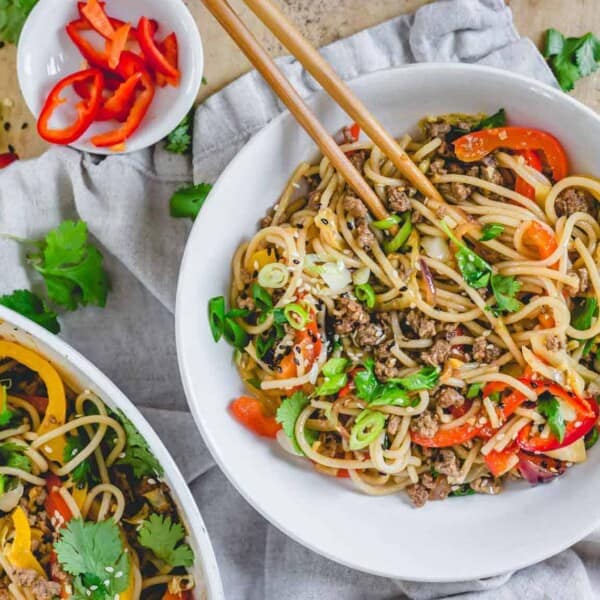 A bowl of stir-fried noodles with beef, red bell peppers, and green onions, garnished with sesame seeds and cilantro. Wooden chopsticks are placed on the bowl. A larger serving bowl with more noodles and a small dish of sliced red bell peppers are in the background.
