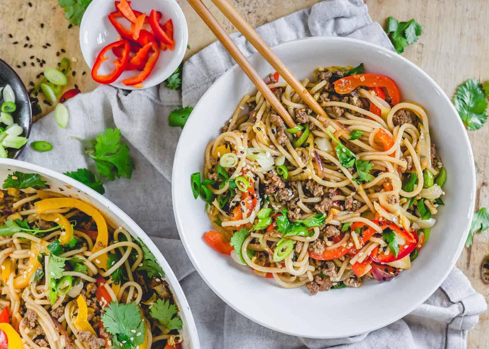 A bowl of stir-fried noodles with beef, red bell peppers, and green onions, garnished with sesame seeds and cilantro. Wooden chopsticks are placed on the bowl. A larger serving bowl with more noodles and a small dish of sliced red bell peppers are in the background.