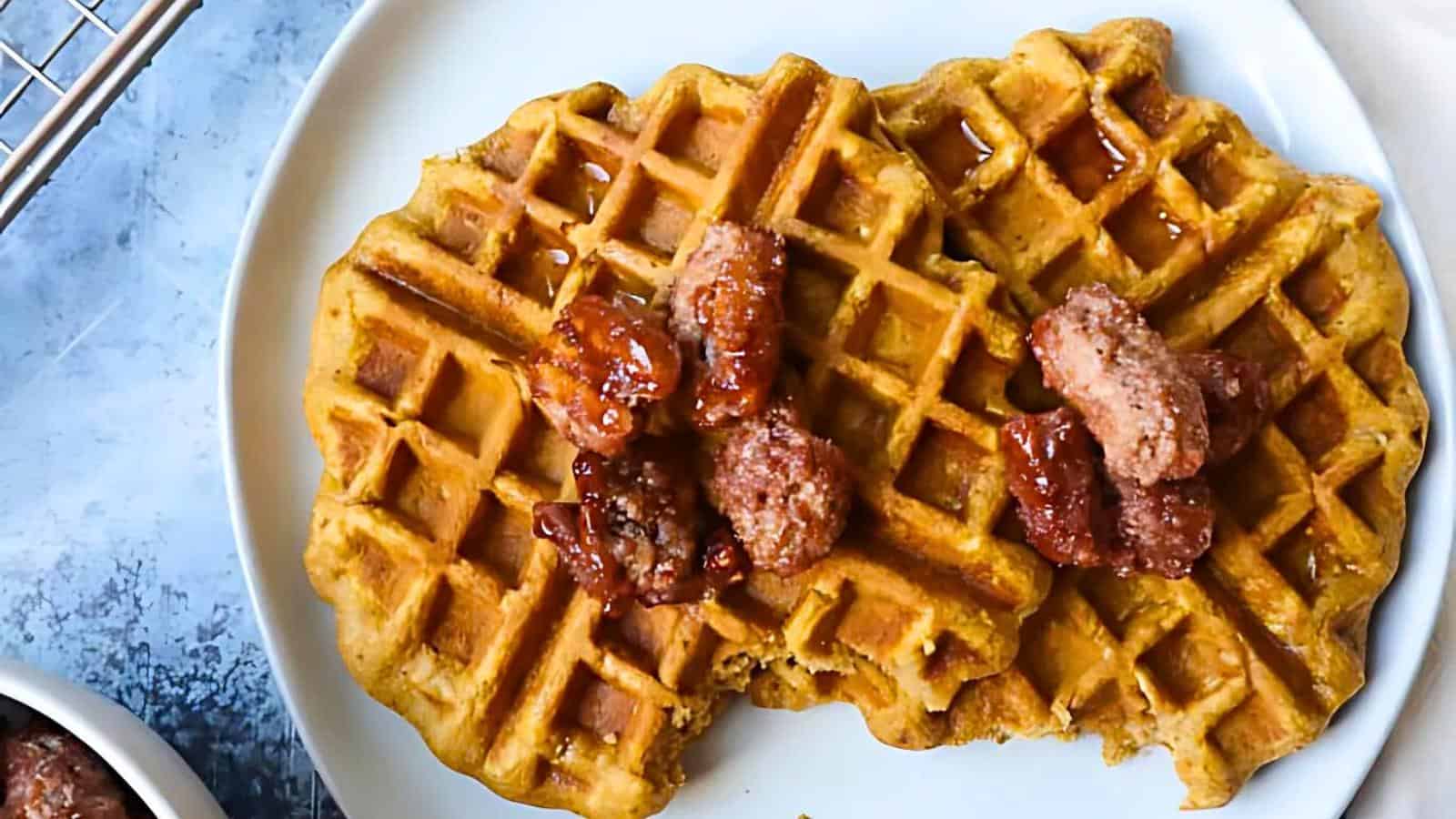 A plate with two golden brown waffles topped with sprinkled candied pecans. The waffles appear crispy and have a slight sheen from a syrup drizzle. The blue background includes part of a cooling rack and a blurred bowl in the lower left corner.