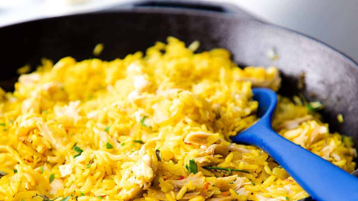 Close-up of a skillet filled with vibrant yellow rice mixed with shredded chicken and garnished with herbs. A blue serving spoon rests in the rice, ready to dish out the meal.