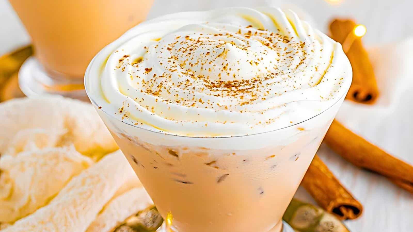 A creamy beverage, light tan in color, served in a martini glass, topped with a generous swirl of whipped cream and a sprinkle of cinnamon. Cinnamon sticks and a white textured cloth are in the background, hinting at a cozy, festive atmosphere.