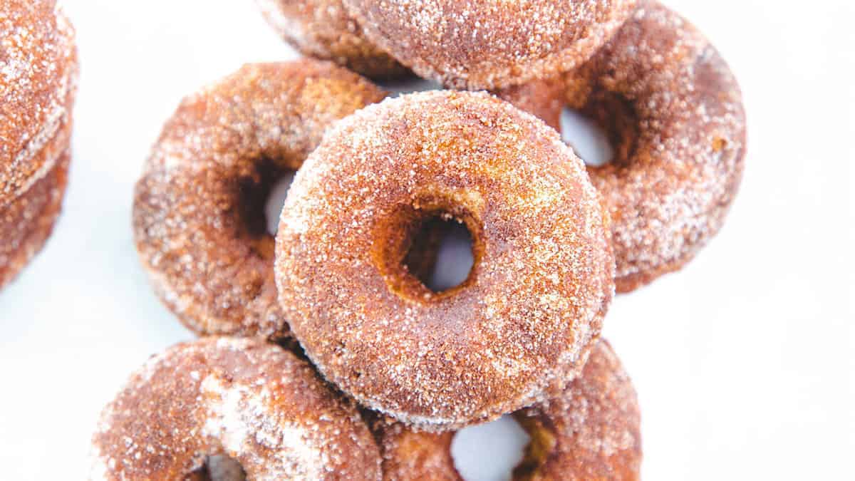 A close-up of several freshly baked, sugar-coated cinnamon donuts stacked on top of each other. The donuts are golden brown with a slightly rough texture from the sugar and cinnamon mixture.