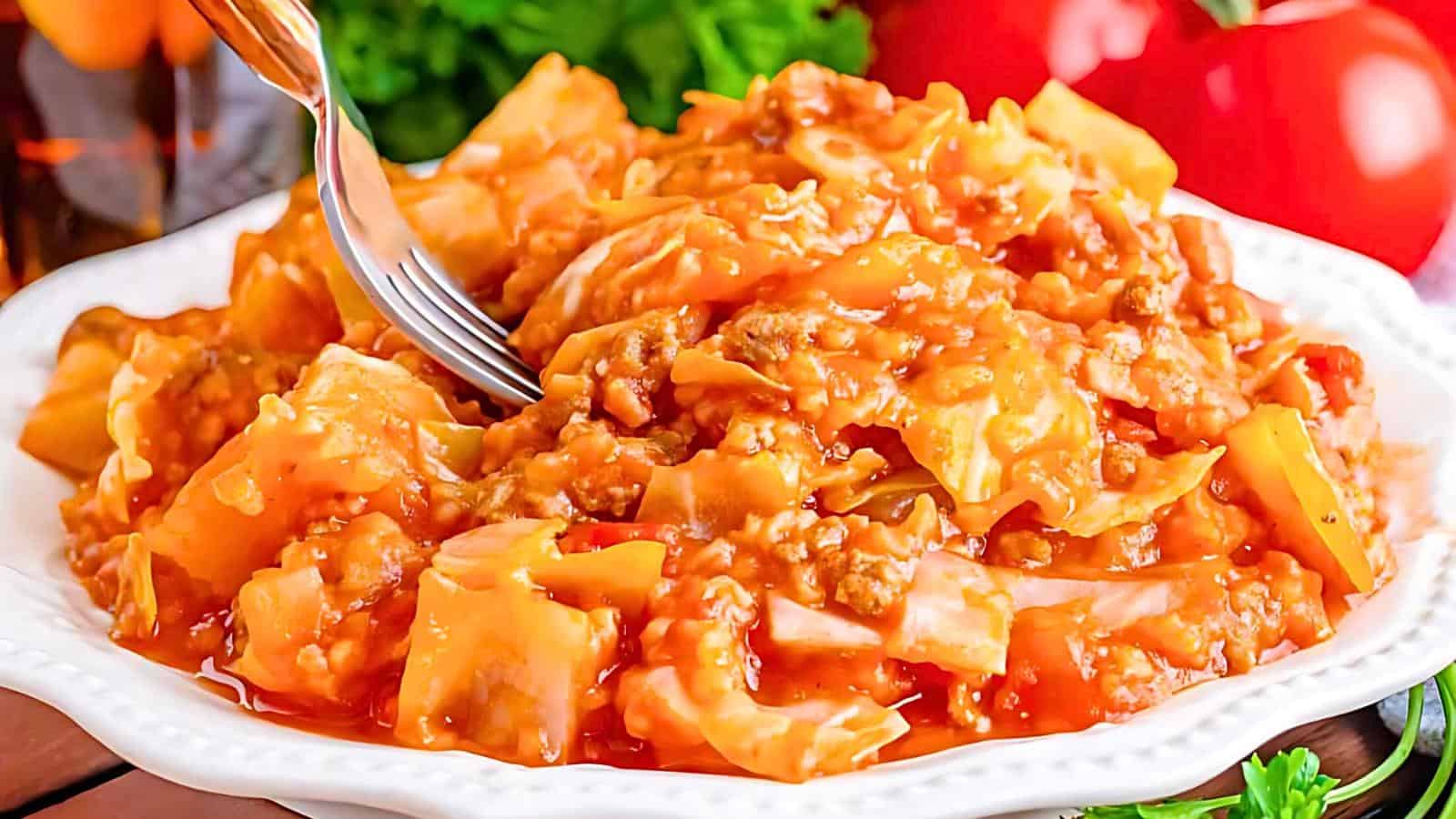 A white plate is filled with a hearty dish of stuffed cabbage casserole, featuring pieces of cabbage, ground meat, and a rich tomato sauce. A fork is poised, ready to take a bite. In the background, fresh ingredients including tomatoes and greens are visible.