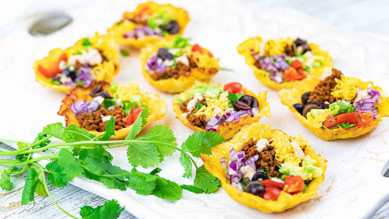 Mini taco bowls filled with ground meat, shredded cheese, cherry tomatoes, black olives, lettuce, and purple cabbage are arranged on a white surface. Fresh cilantro is placed nearby as garnish. The taco bowls appear crispy and golden.