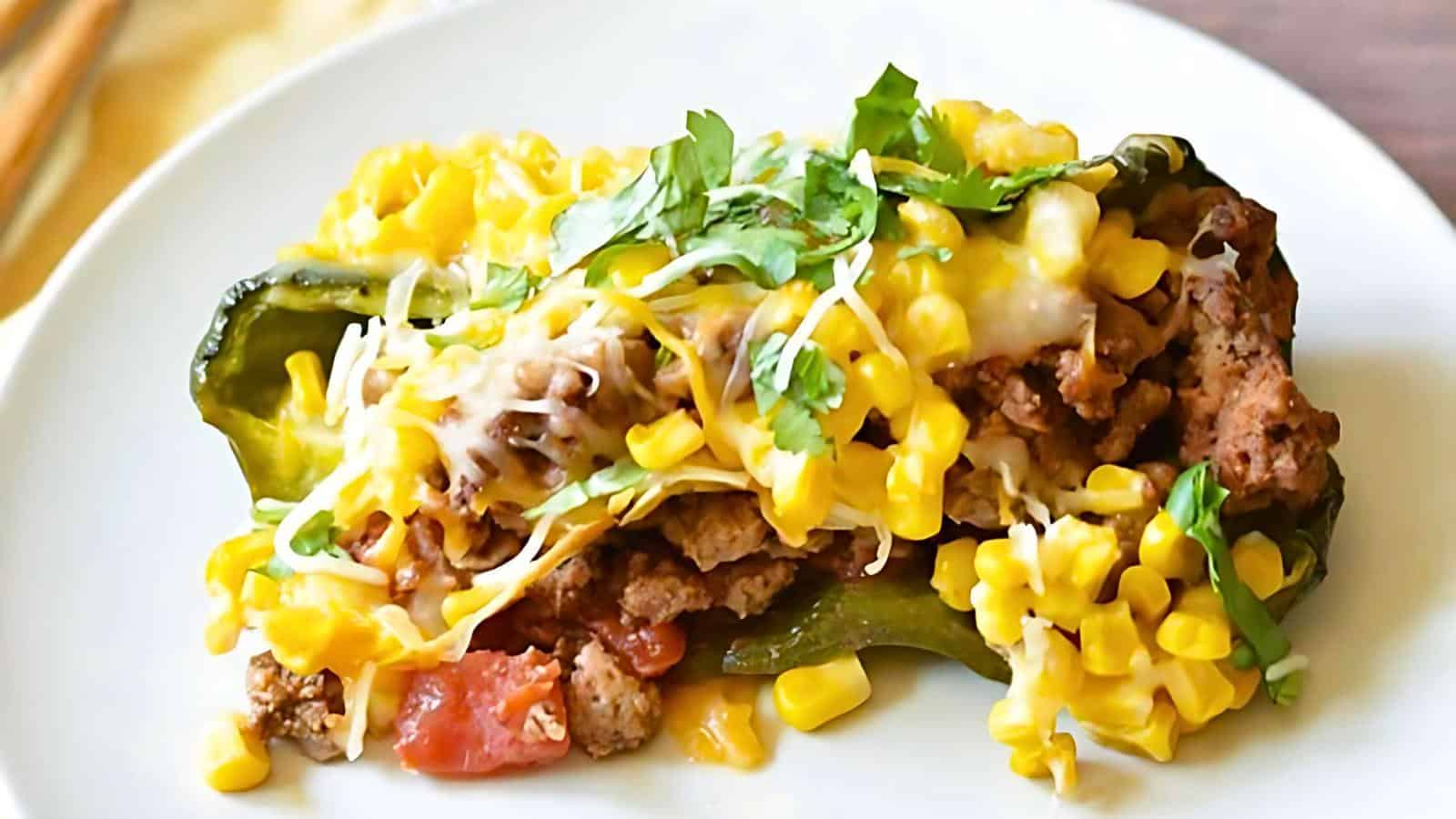 A dish featuring a stuffed roasted poblano pepper filled with ground meat, tomatoes, and topped with melted cheese, fresh corn kernels, and chopped cilantro, served on a white plate.