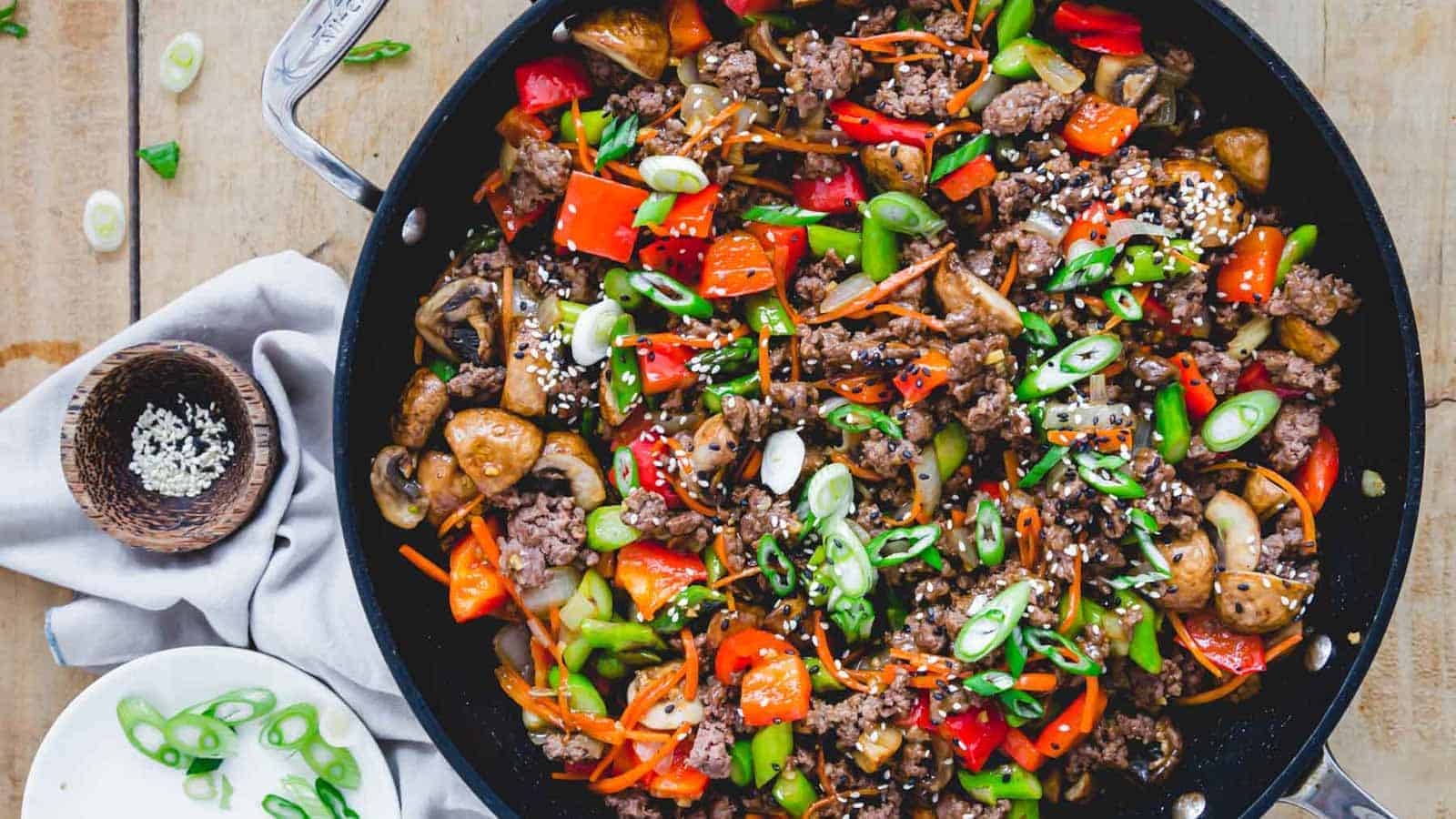 A close-up of a skillet containing a vibrant Asian stir-fry dish with ground beef, red and green bell peppers, mushrooms, and sliced green onions. The dish is garnished with sesame seeds. Nearby are a small bowl of extra sesame seeds and a plate with additional chopped green onions.